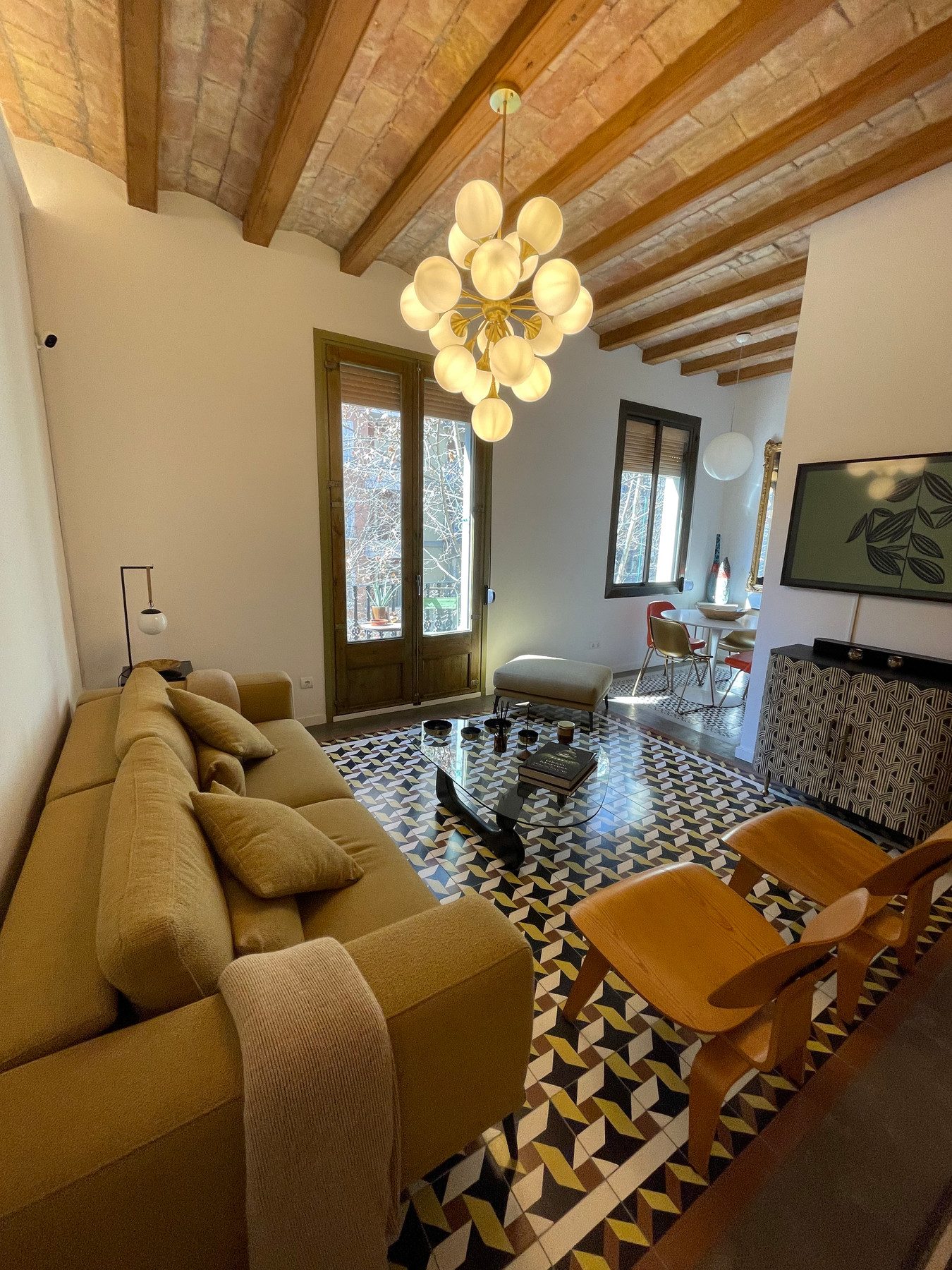 Salón con techo de vigas de madera, suelo hidráulico y sofá mostaza en una reforma integral en Carrer Viladomat, Barcelona